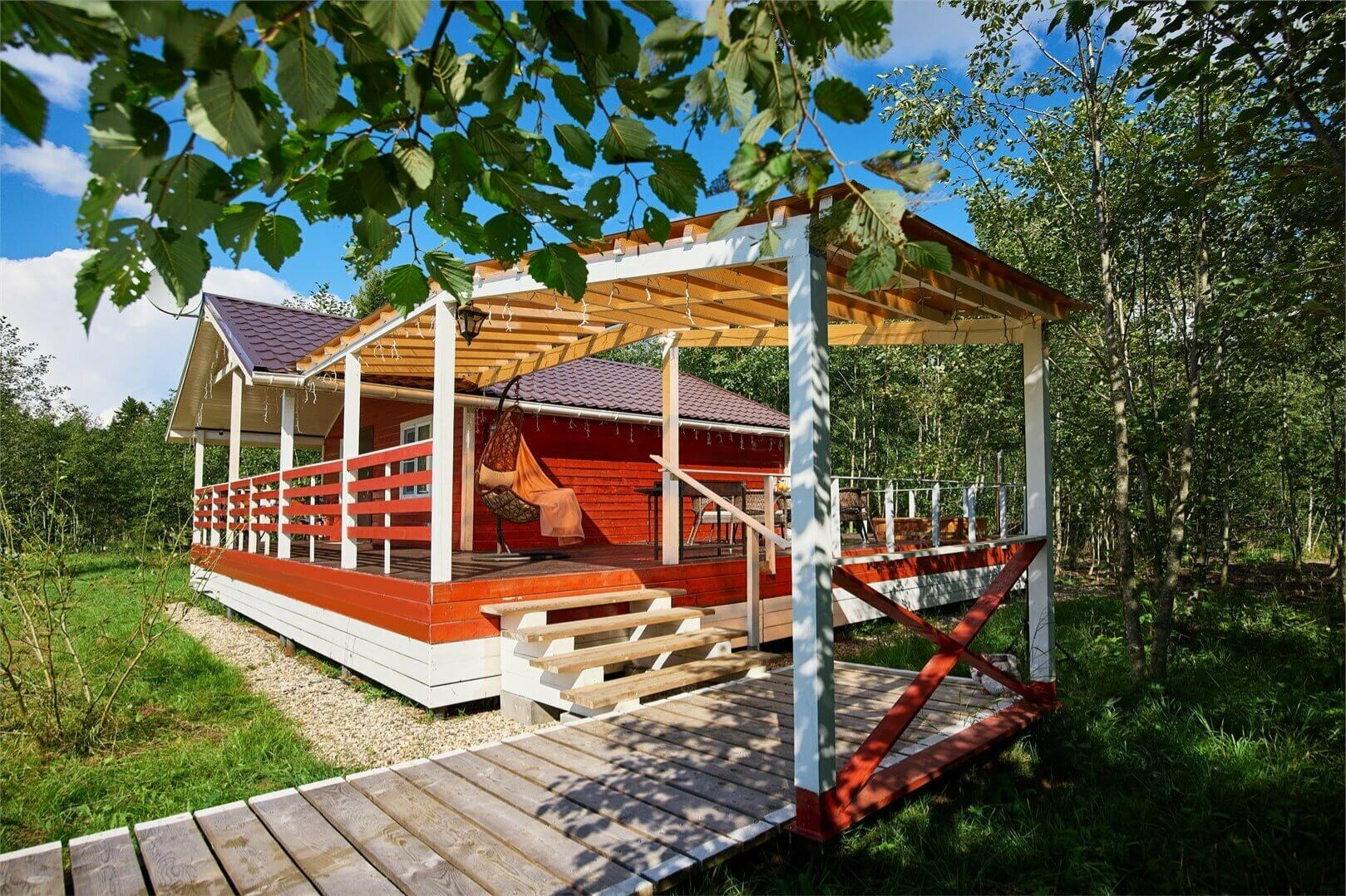 Красная веранда с перголой среди берёз и хвойного леса.