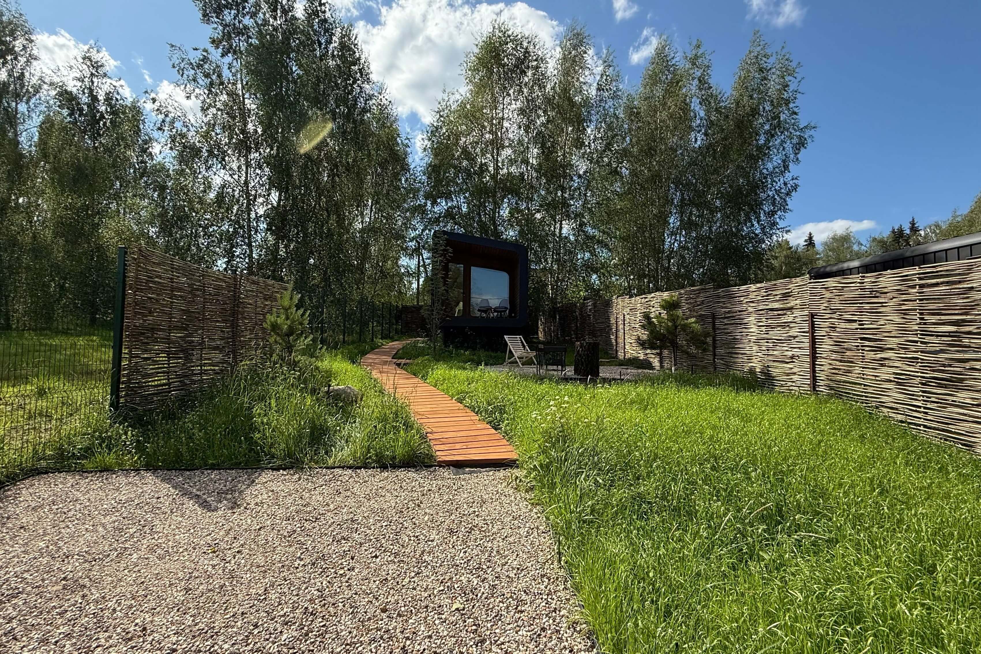 Дорожка между плетёными заборами к чёрному срубу среди берёз.