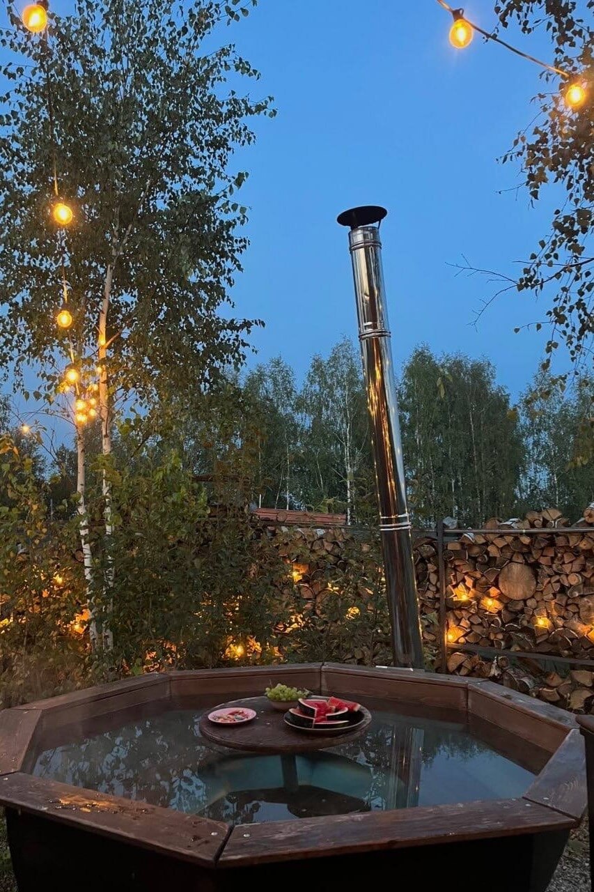 Деревянная купель с трубой-дымоходом под березами в сумерках.