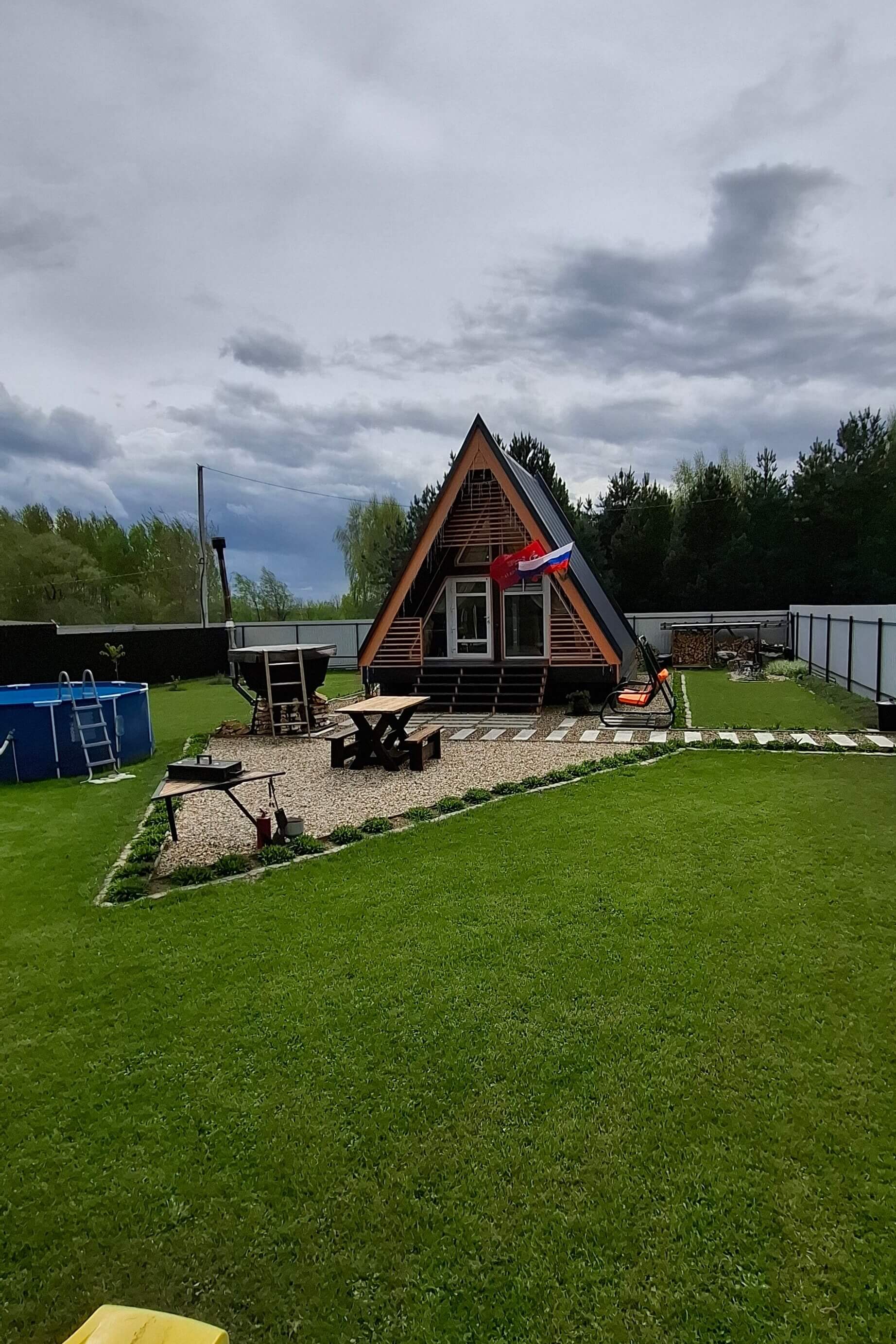 А-образный домик с красной крышей на берегу озера среди сосен.