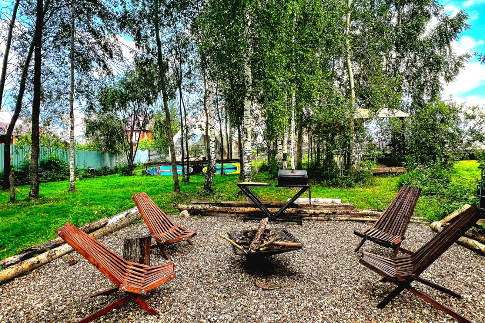 Костровая площадка с лежаками среди берёз и зелени.